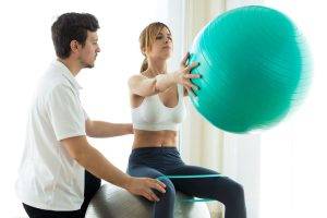 Shot of physiotherapist helping patient to do exercise on fitness ball in physio room.